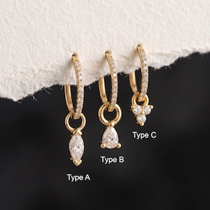 May include: Three gold hoop earrings with clear stone accents. Type A features a marquise-cut stone charm, Type B has a pear-cut stone charm, and Type C displays a three-stone cluster charm. Each hoop is pavé-set with small clear stones.