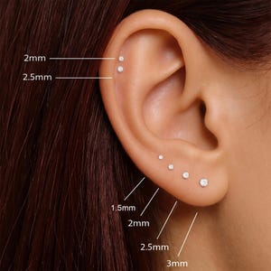 Può includere: Un set di sei piccoli orecchini a lobo rotondi e trasparenti disposti lungo il lobo e la cartilagine. Gli orecchini sono etichettati con le misure 1,5 mm, 2 mm, 2,5 mm e 3 mm, che indicano il diametro di ogni singola pietra incastonata.