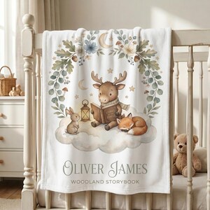 May include: A white fleece blanket featuring a central illustration of a deer reading a book titled Stories of the Forest, a rabbit, and a sleeping fox on a cloud. The design includes a floral wreath, stars, and the text Oliver James Woodland Storybook.