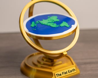 Könnte beinhalten: Ein goldfarbenes, dekoratives Flachweltmodell. Das Modell zeigt eine blaue Scheibe mit grünen Kontinenten, eingerahmt von einem goldenen Ring und Sockel. Der Sockel trägt den Text "The Flat Earth".