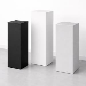 May include: Three rectangular pedestals in a studio setting. One is black, the other two are white. The pedestals are of varying heights and widths, and are placed on a light gray floor against a white wall.