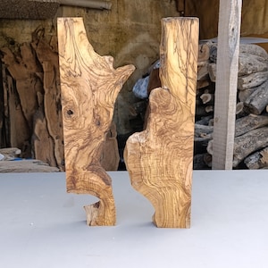 Pode incluir: Duas placas verticais de madeira de oliveira natural com padrões de veios castanhos escuros e ondulados sobre um fundo castanho claro. Cada peça apresenta bordos orgânicos irregulares e uma superfície lisa com nós visíveis.