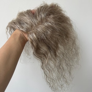 May include: A hair topper featuring wavy, light brown and grey highlighted strands. The piece has a visible centre part and a natural texture, showcasing a blend of cool-toned ash brown and silver-grey hair fibres arranged in a layered style.