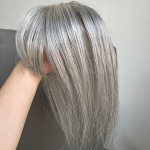 Op de afbeelding: Een haartopper met steil, schouderlang haar in een mix van zilveren, grijze en witte tinten. Het haarstuk is voorzien van een zijwaartse pony en een zichtbare kanten basis bij de kruin, ontworpen voor een natuurlijk ogende dekking.