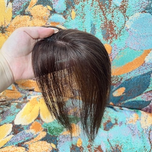 May include: A dark brown hair topper with a fringe. The hairpiece is held up against a colorful floral patterned background. The hair appears to be human hair.