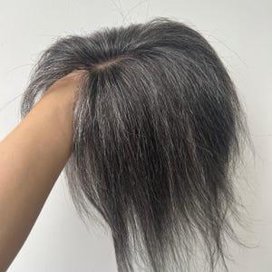 Puede incluir: Una pieza de cabello gris y blanco, sostenida por una mano. La pieza capilar tiene un aspecto natural con diferentes tonos de gris y blanco, y parece estar hecha de cabello humano. El cabello está peinado con una ligera onda.