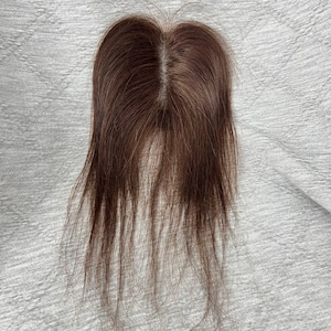 May include: A brown hair topper with a center part, designed to add volume and coverage. The hairpiece is a warm brown color and appears to be made of natural hair. The topper is placed on a white, textured surface.