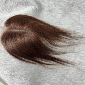 Op de afbeelding: Een bruin haarstuk met een middenscheiding. Het haarstuk heeft een roodbruine kleur en lijkt van echt haar te zijn gemaakt. Het haar is glad en steil, met een natuurlijke glans. Het haarstuk is ontworpen om volume toe te voegen.