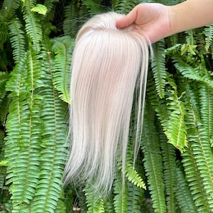 Puede incluir: Un postizo de cabello con mechones lisos de color rubio platino y una base de monofilamento visible en la coronilla. El cabello es liso y sedoso, mostrando un color rubio claro de tonos fríos en toda la longitud de la pieza.
