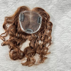 May include: A brown curly hair topper with a mesh base and three clips for secure attachment. The hairpiece is designed to add volume and coverage to the top of the head. The hair is a warm, reddish-brown color.