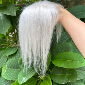 May include: A hair topper piece featuring straight, long, silver-white hair strands. The hair is styled with a center part and has a smooth, sleek texture that flows downward. The hairpiece is designed to provide coverage and volume for hair.