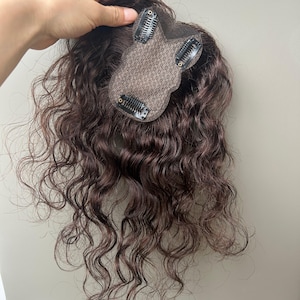 Könnte beinhalten: Ein dunkelbraunes Haarteil mit welligem, lockigem Haar. Das Haarteil hat eine kleine, braune Mesh-Basis mit vier Clips zur Befestigung. Das Haarteil ist so konzipiert, dass es dem natürlichen Haar Volumen und Deckkraft verleiht.