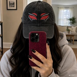 Peut inclure: Une casquette de baseball en coton délavé gris anthracite avec un motif brodé représentant deux yeux stylisés rouges et argentés sur le panneau avant. La casquette présente une visière incurvée et une calotte structurée avec des coutures visibles.