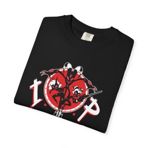 May include: A black t-shirt featuring a graphic print of three animated characters in red, black, and white. The design includes a large red circle with white dots, the letters I.M.P in white, and characters holding a rifle and other accessories.