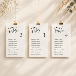 Puede incluir: Tres tarjetas de mesa rectangulares blancas colgadas de cuerdas finas con clips de metal dorado. Cada tarjeta presenta el texto negro Table seguido de un número 2, 3 o 4, y una lista de nombres que incluye a Abigail Cooper y Michael Parker.