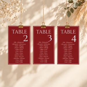 Puede incluir: Tres tarjetas de mesa rectangulares de color rojo intenso colgadas de clips dorados. Cada tarjeta presenta el texto blanco TABLE seguido de los números 2, 3 o 4, y una lista de nombres en fuente cursiva blanca, con un borde blanco fino.
