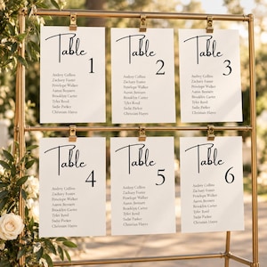 Può includere: Un supporto in metallo dorato che sostiene sei cartoncini rettangolari bianchi con testo nero. Ogni cartoncino riporta la parola Table seguita da un numero da 1 a 6 e un elenco di nomi tra cui Audrey Collins, Zachary Foster e Penelope Walker.
