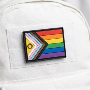 May include: A rectangular patch featuring a rainbow flag design with a black border. The patch includes the colors of the LGBTQ+ pride flag, as well as the colors of the intersex flag and the transgender flag. The patch is affixed to a white fabric surface.