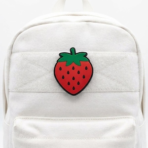 May include: A white backpack with a red and green strawberry patch. The strawberry patch is in the center of the backpack, with a red body, green stem, and black seeds. The backpack has a front pocket and a top handle.