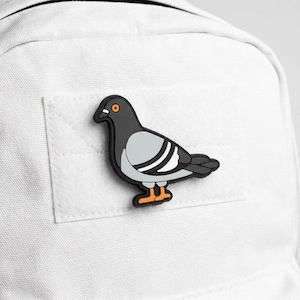 May include: A black, gray, and white pigeon-shaped patch with orange feet and eye. The patch is affixed to a white backpack. The pigeon has a detailed design, with a gray wing and a black outline.