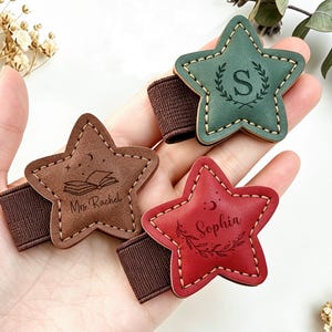 Puede incluir: Tres pinzas para el pelo en forma de estrella de cuero marrón, verde y rojo. Cada pinza tiene un diseño diferente: un libro y el nombre "Mrs. Rachel", una letra "S" dentro de una corona y el nombre "Sophia" con un adorno floral. Todas tienen bandas elásticas marrones.