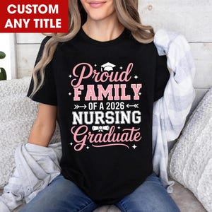 May include: A black short-sleeved t-shirt featuring pink and white text that reads Proud FAMILY OF A 2026 NURSING Graduate. The design includes a graphic of a graduation cap above the word Proud and a diploma scroll icon above the word Graduate.