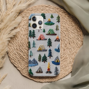 May include: A light gray phone case with an embroidered camping scene. The design includes colorful tents, trees, mountains, and campfires. The case has a textured appearance and a nature-inspired theme.