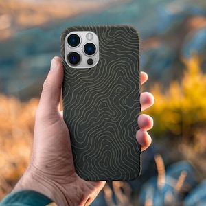 May include: A black phone case with a topographic map design in gold lines. The case is held up in front of a blurred natural background. The phone case has multiple camera lenses.