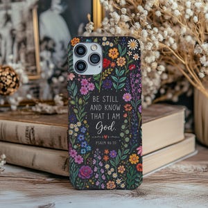 May include: A black phone case adorned with a vibrant floral design in shades of pink, purple, orange, and yellow. The case features the text "BE STILL AND KNOW THAT I AM God." and "PSALM 46:10". The case is surrounded by books and flowers.
