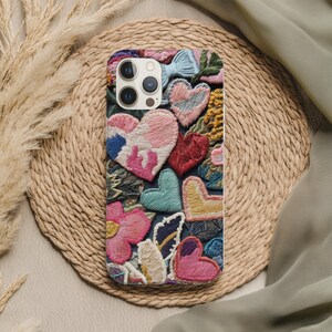 May include: Embroidered phone case featuring a colorful design of hearts and flowers in shades of pink, red, blue, and yellow. The case has a textured appearance with detailed stitching, creating a unique and artistic look.