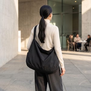 May include: A large, black shoulder bag with a rounded shape. The bag is worn over the shoulder. The person is wearing a cream-colored turtleneck sweater and dark gray pants. The background includes a building and people sitting at a table.