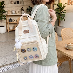May include: A white backpack featuring a top handle, adjustable shoulder straps, and a front pocket with a mesh overlay. The bag displays a brown patch with the text XDTU FASHION, a white plush bear keychain, and five circular pins attached to the mesh.