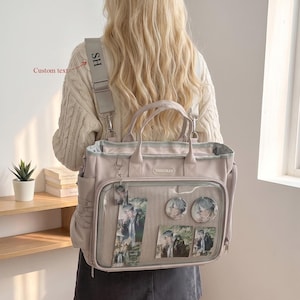 May include: A light beige tote bag with a clear front display pocket containing anime-style character prints and circular pins. The bag features top handles, a detachable shoulder strap with the text HS, and the brand name YISHUIRAN on the front.