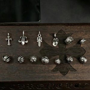 May include: A selection of silver pendants, rings, and beads arranged on a dark wooden surface. The pendants showcase diverse designs, such as a cross, fleur-de-lis, and a stylized dagger. The beads have a ribbed pattern.