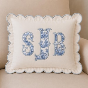 May include: A rectangular off-white throw pillow featuring a scalloped edge with light blue piping. The center displays a large embroidered monogram with the letters SJB, filled with a blue and white toile pattern depicting pastoral scenes.