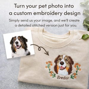May include: A beige sweatshirt with a custom embroidered dog portrait. The embroidery features a brown, white, and black dog with orange floral accents and the name "Eredar" below. The image also shows a photo of the dog.