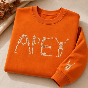 May include: An orange crewneck sweatshirt featuring an embroidered design of skeletons forming the letters A, P, E, and Y across the chest. The right sleeve cuff displays an additional embroidered skeleton hand making a rock and roll gesture.