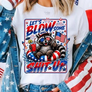 Könnte beinhalten: Ein weißes T-Shirt mit einer Grafik eines Waschbären, der eine Sonnenbrille und eine Mütze trägt und in einem patriotischen Schwimmring sitzt. Der Waschbär hält ein Getränk und eine Flagge. Der Text lautet LET'S BLOW SOME SHIT UP mit Feuerwerk.