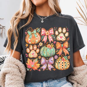 Puede incluir: Una camiseta gris carbón de manga corta con un estampado gráfico colorido de tres calabazas, tres lazos, dos huellas de pata y una hoja de arce. Las calabazas y las huellas tienen motivos florales y diseños de cuadros en naranja, turquesa y morado.