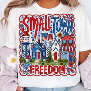 Könnte beinhalten: Weißes T-Shirt mit einem farbenfrohen Grafikdesign mit der Aufschrift "SMALL TOWN FREEDOM". Das Design zeigt einen Wasserturm, Häuser, eine amerikanische Flagge und eine Kirche im patriotischen Stil.