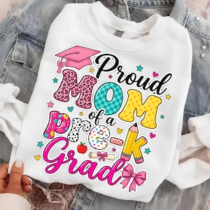 Può includere: Felpa bianca con la scritta "Proud Mom of a Pre-K Grad" in un design colorato. Il design include un cappello di laurea, stelle, cuori e una matita. Le lettere sono decorate con motivi.