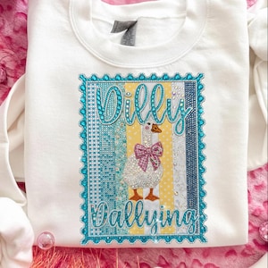 May include: White sweatshirt featuring a rhinestone design with the words "Dilly Dallying" and a goose wearing a pink bow. The design is framed with a turquoise border and includes yellow and blue stripes.