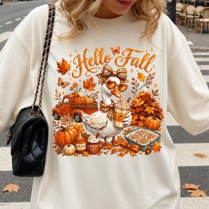 Puede incluir: Una sudadera color crema con el gráfico de un ganso que lleva gafas en forma de corazón y un lazo a cuadros, sosteniendo un café. El diseño incluye el texto 'Hello Fall', un camión naranja, calabazas, hojas de otoño y rollos de canela.