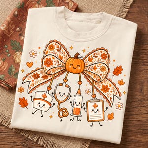 May include: A cream-colored t-shirt featuring a large graphic of a pumpkin-centered bow with floral patterns. Dangling from the bow are cartoon-style medical items including a nurse cap, stethoscope, syringe, and clipboard, all with smiling faces.