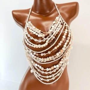 May include: A halter-style body chain top constructed from multiple draped strands of white and cream-colored circular shell discs and small round beads. The piece features a layered design that hangs across the chest, secured by a thin neck strap.