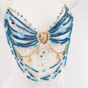 Pu&ograve; includere: Un top all'americana realizzato con molteplici fili di perline di cristallo blu e trasparenti. Il design presenta catene in metallo dorato, piccoli ciondoli a forma di conchiglia dorata e conchiglie blu iridescenti attorno a una conchiglia.