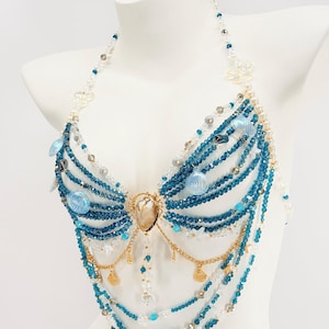 Pu&ograve; includere: Un top di perline stile allacciato al collo con fili di cristalli blu e trasparenti, perle bianche e ciondoli a forma di conchiglia azzurra. Il design include catene in metallo dorato con piccoli pendenti a conchiglia e un cuore centrale iridescente.