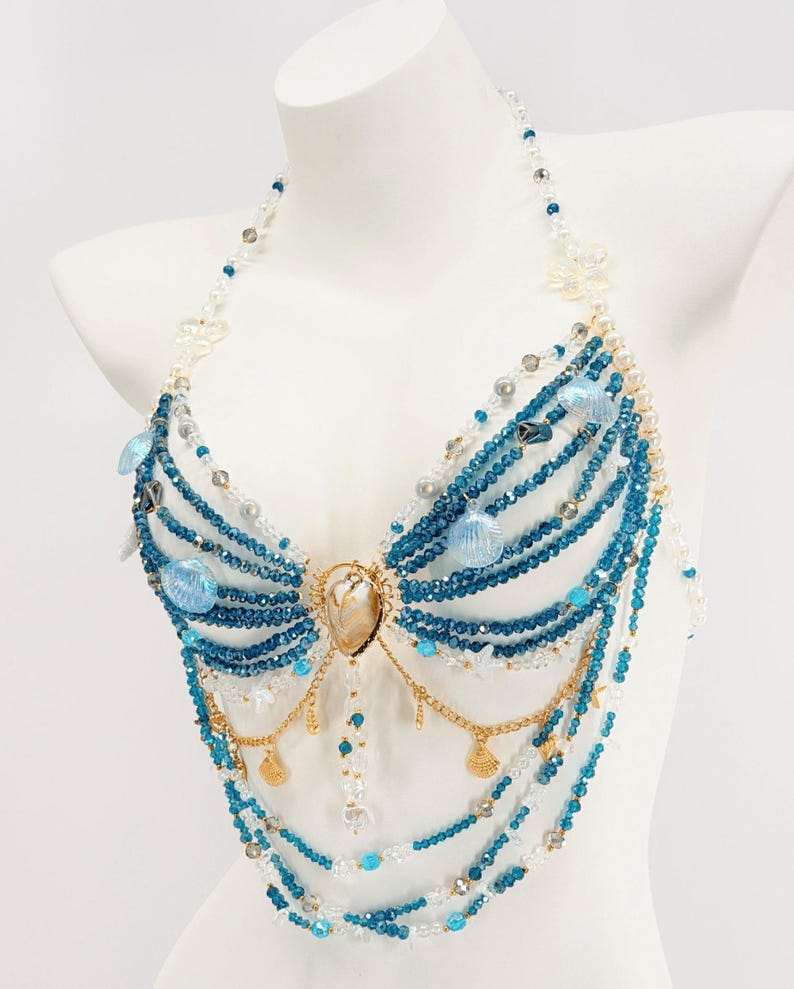 Pu&ograve; includere: Un top di perline stile allacciato al collo con fili di cristalli blu e trasparenti, perle bianche e ciondoli a forma di conchiglia azzurra. Il design include catene in metallo dorato con piccoli pendenti a conchiglia e un cuore centrale iridescente.
