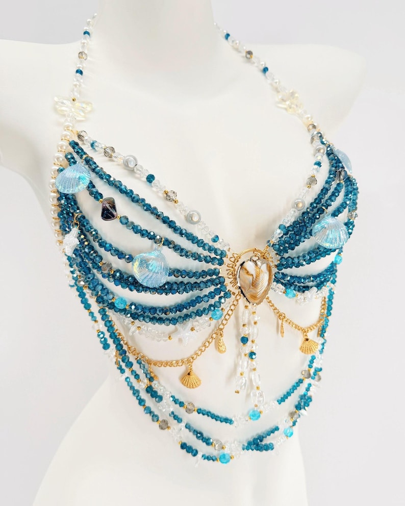 Pu&ograve; includere: Una catena per il corpo stile halter con molteplici fili di perline di cristallo blu profondo e trasparenti. Il design incorpora catene in metallo dorato, piccoli ciondoli a forma di conchiglia dorata e conchiglie blu iridescenti.