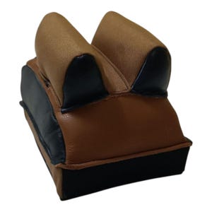 Puede incluir: Un soporte de tiro marrón y negro con forma de silla de montar. El soporte presenta una base de cuero marrón, detalles de cuero negro y una parte superior de tela beige. El diseño está pensado para proporcionar estabilidad a las armas de fuego durante la práctica de tiro o la caza.